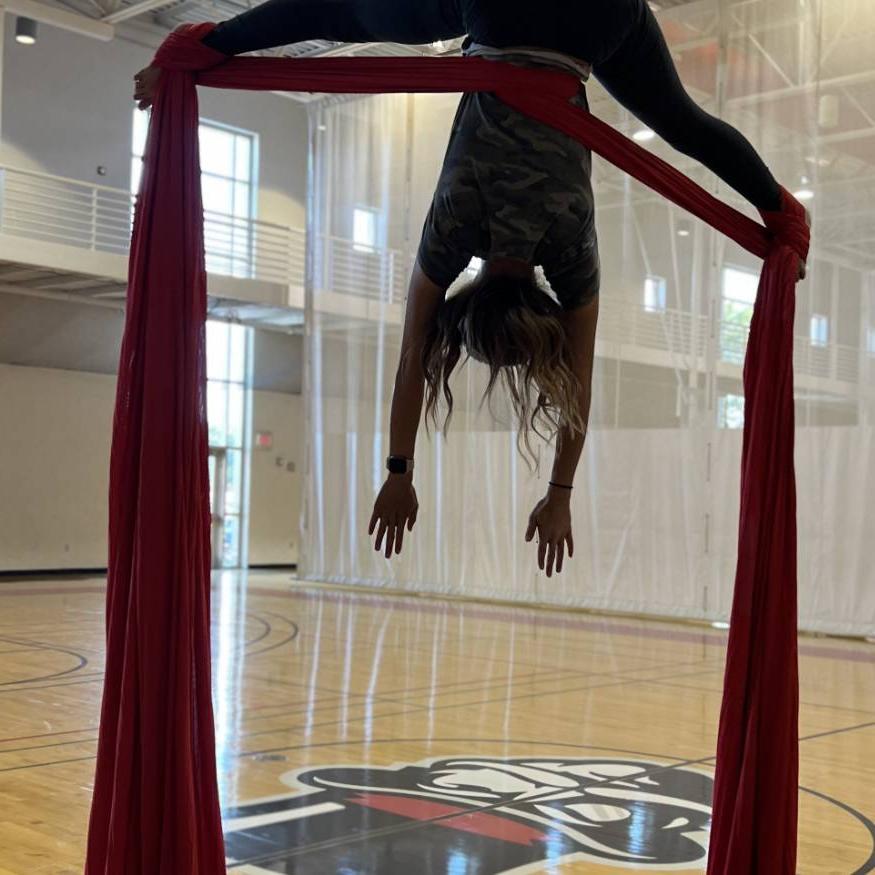 Aerial Silks student on red silk in crossback straddle, with gov head in background on gym court floor